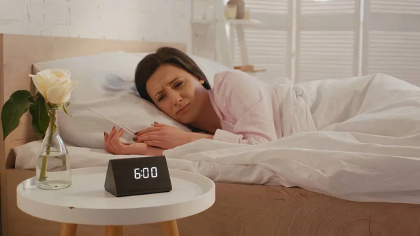 Upset woman looking at clock on bedside table in bedroom — стоковое фото