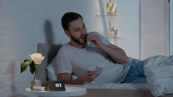 Young man eating chocolate bar on bed at night — Stockfoto