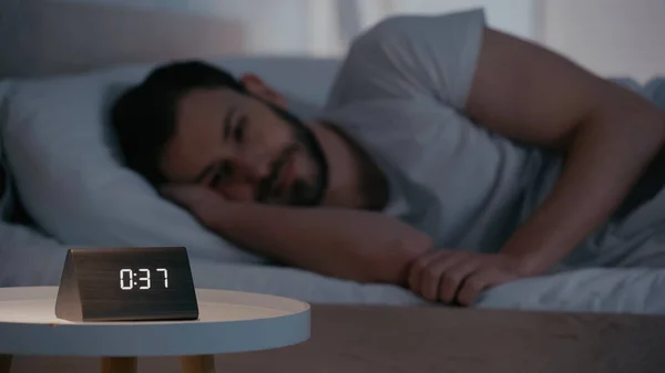 Clock on bedside table near blurred man lying on bed at night — Stock Photo