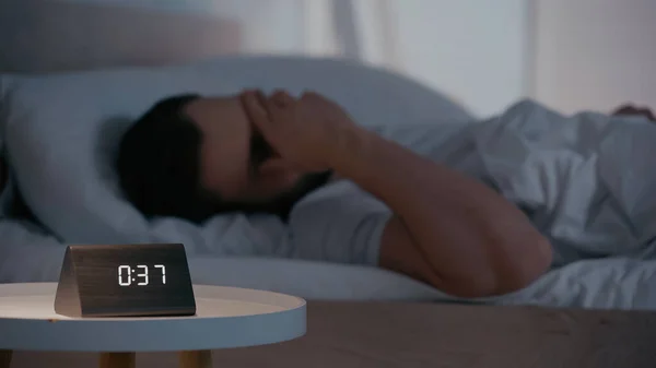 Clock on bedside table near blurred man covering face on bed at night — Stock Photo