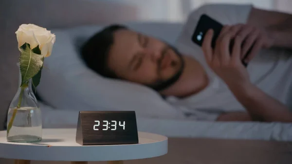 Planta y reloj en la cama cerca del hombre borroso utilizando el teléfono inteligente en la cama en casa - foto de stock