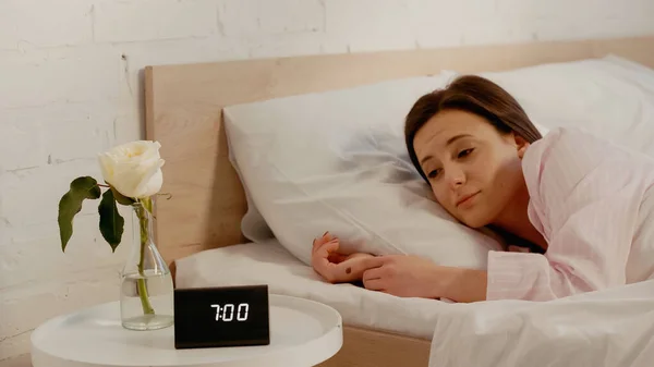 Sleepy woman looking at clock near flower in bedroom — Stockfoto