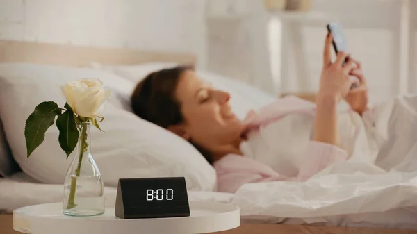 Clock near rose in vase and blurred woman using smartphone on bed — Fotografia de Stock