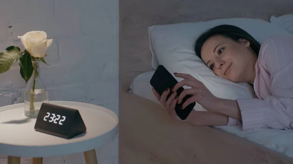 Smiling woman using smartphone on bed at night — стоковое фото