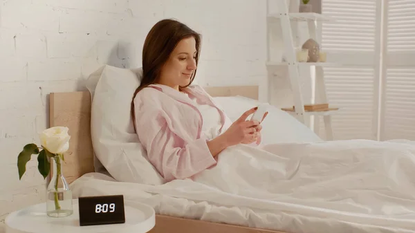 Счастливая женщина использует смартфон на кровати рядом с часами и цветок на тумбочке — стоковое фото
