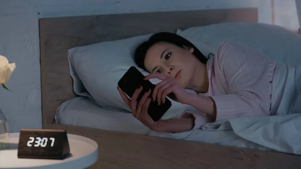 Brunette woman in pajamas using smartphone near blurred clock in bedroom — Foto stock