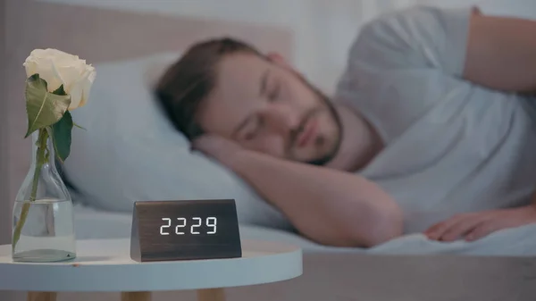 Rose and clock on bedside table near sleeping man at home — Stock Photo