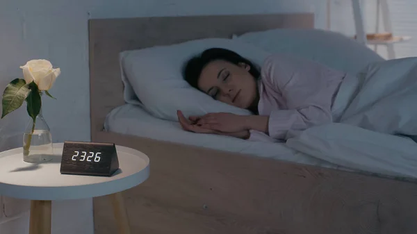 Fleur et horloge sur la table de chevet près de la femme dormant à la maison — Photo de stock