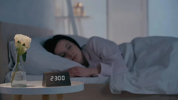 Flower and clock on bedside table near blurred brunette woman sleeping at home — Fotografia de Stock