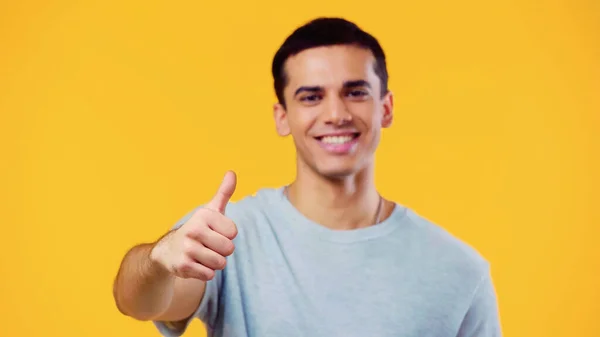 Sonriente y borroso hombre mirando a la cámara mientras muestra el pulgar hacia arriba aislado en amarillo - foto de stock