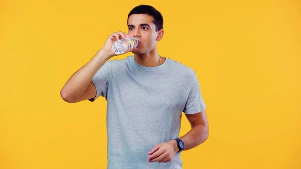 Young man in t-shirt holding bottle while drinking water isolated on yellow — Stock Photo