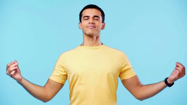 Pleased young man with closed eyes in yellow t-shirt meditating isolated on blue — Stock Photo