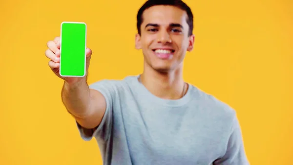 Blurred and cheerful young man in t-shirt showing smartphone with green screen isolated on yellow — Stock Photo