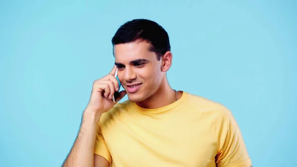 Smiling young man in yellow t-shirt talking on cellphone isolated on blue — Stock Photo