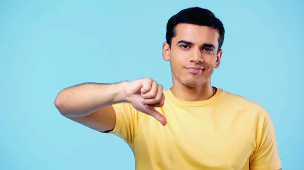 Displeased young man in yellow t-shirt showing thumb down isolated on blue — Fotografia de Stock