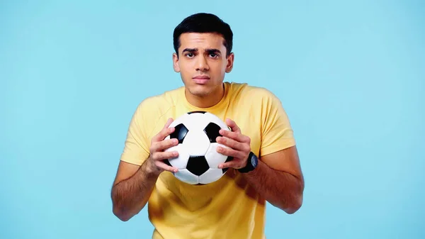 Jovem estressado em camiseta amarela segurando futebol isolado em azul — Fotografia de Stock