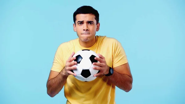 Joven preocupado en camiseta amarilla sosteniendo el fútbol aislado en azul - foto de stock