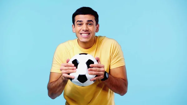 Feliz joven en camiseta amarilla sosteniendo el fútbol aislado en azul - foto de stock