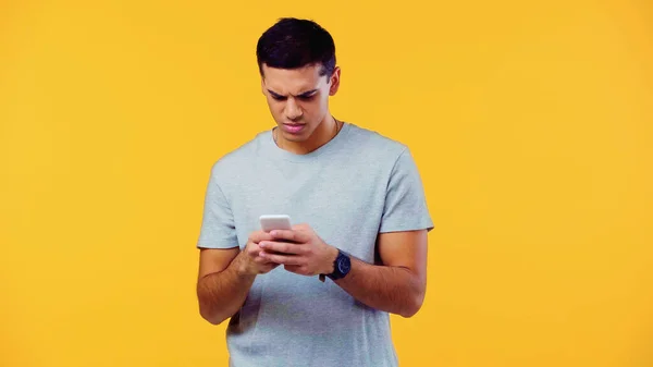 Сумний молодий чоловік у футболці обміну повідомленнями мобільний телефон ізольований на жовтому — стокове фото