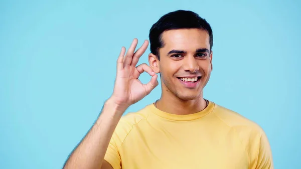 Cheerful young man in yellow t-shirt showing ok sign isolated on blue — Fotografia de Stock