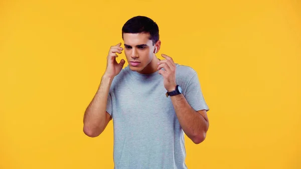 Спантеличений молодий чоловік у футболці, що регулює бездротові навушники ізольовані на жовтому — стокове фото