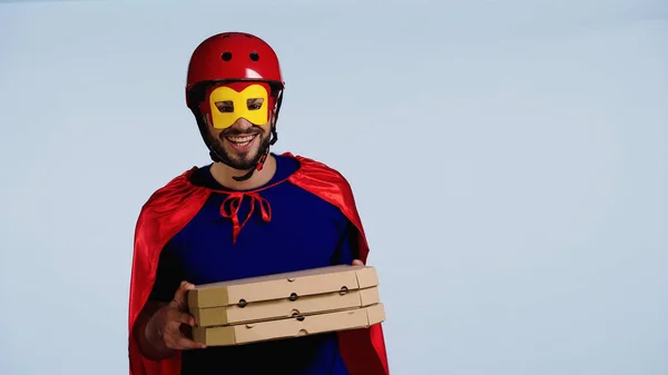 Happy man in superhero costume holding boxes with pizza isolated on blue — Stock Photo