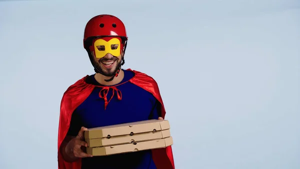 Happy man in superhero costume holding boxes with pizza isolated on blue — стоковое фото