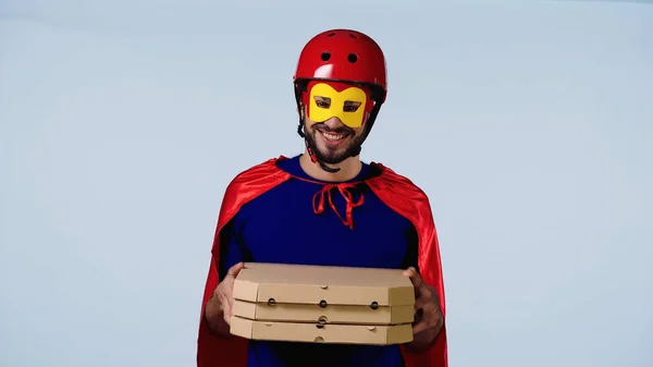 Smiling man in superhero costume holding boxes with pizza isolated on blue — стоковое фото