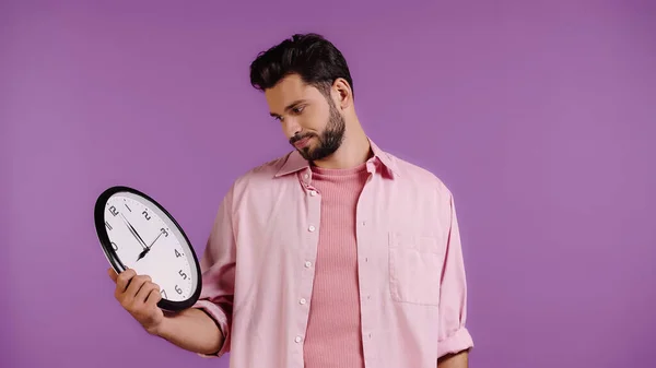 Displeased young man looking at clock isolated on purple — Foto stock