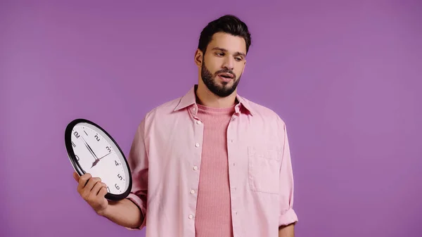 Dissatisfied young man holding clock isolated on purple — Stock Photo