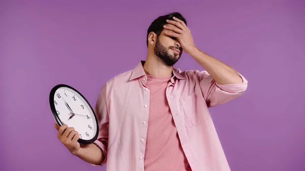 Обеспокоенный молодой человек держит часы и показывая лицо ладони изолированы на фиолетовый — стоковое фото