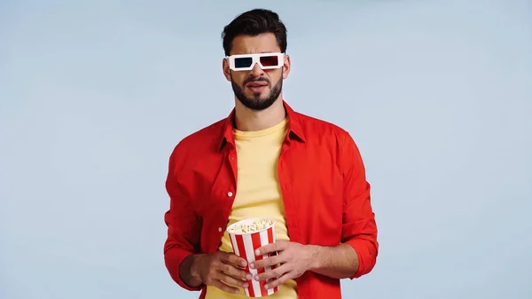 Unzufriedener und bärtiger Mann mit 3D-Brille, der sich einen Film ansieht und Popcorn-Eimer isoliert auf blauem Grund hält — Stockfoto