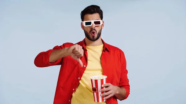 Joven barbudo hombre en gafas 3d viendo la película, mostrando el pulgar hacia abajo y sosteniendo palomitas de maíz cubo aislado en azul - foto de stock