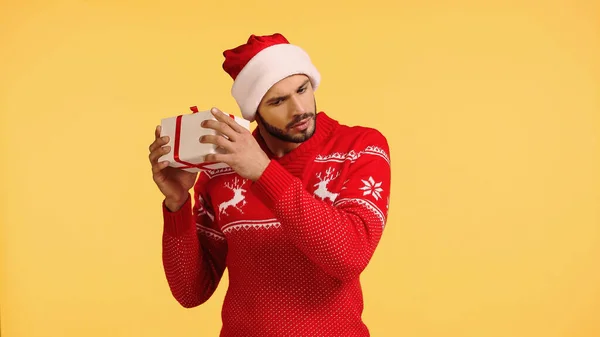 Neugieriger Mann mit Weihnachtsmannhut schüttelt Geschenkbox isoliert auf gelb — Stockfoto