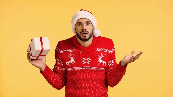 Confuso uomo in cappello Babbo Natale contenente scatola regalo isolato su giallo — Foto stock