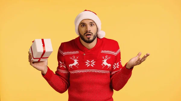 Confused man in santa hat holding gift box isolated on yellow — стоковое фото