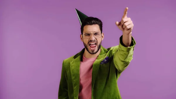 Bearded man in green jacket and party cap pointing with finger and screaming isolated on purple — Stockfoto