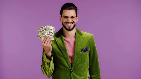 Homem feliz em casaco verde e óculos segurando dólares isolados em roxo — Fotografia de Stock