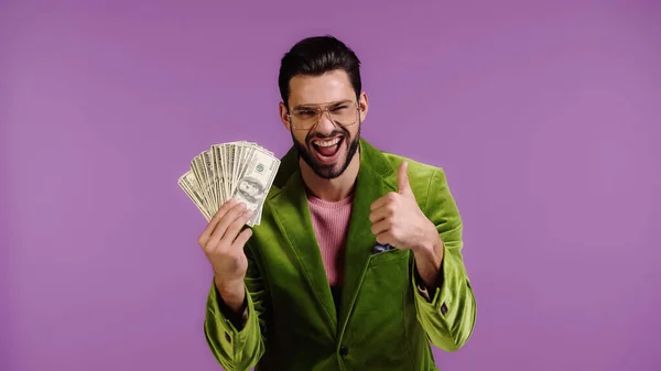 Homem alegre em casaco verde e óculos segurando dólares e mostrando polegar para cima isolado em roxo — Fotografia de Stock