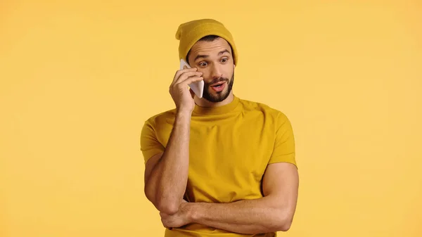 Surprised man in beanie hat talking on smartphone isolated on yellow — стоковое фото