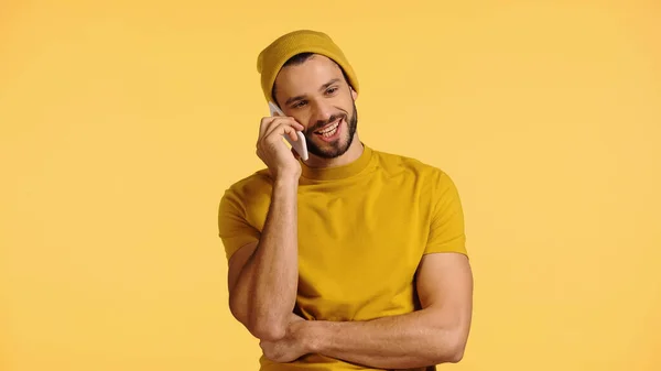 Young man in beanie hat and t-shirt talking on smartphone isolated on yellow — Stock Photo