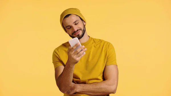 Hombre joven en gorro sombrero y camiseta mirando teléfono inteligente aislado en amarillo - foto de stock