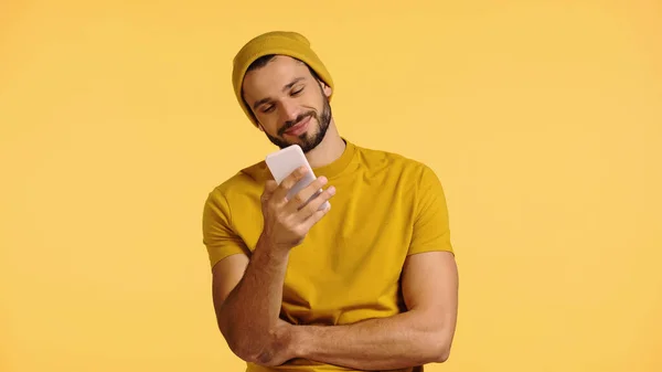 Hombre joven en gorro sombrero y camiseta mirando teléfono inteligente aislado en amarillo — Stock Photo