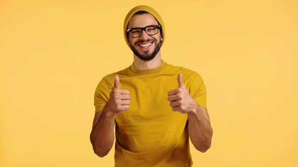 Joven feliz en gorro sombrero, gafas y camiseta que muestra los pulgares hacia arriba aislado en amarillo - foto de stock