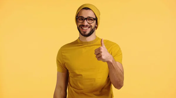 Joven feliz en gorro sombrero, gafas y camiseta que muestra el pulgar hacia arriba aislado en amarillo - foto de stock