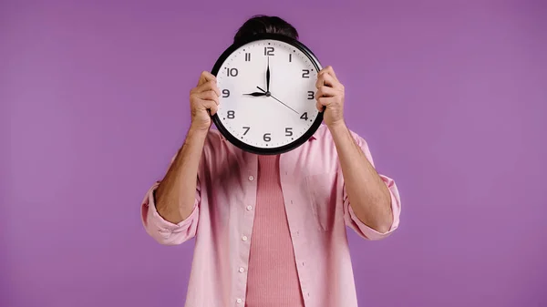 Jeune homme en chemise rose visage obscurcissant avec horloge isolé sur violet — Photo de stock