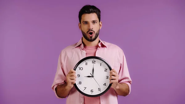 Chocado jovem homem segurando relógio isolado em roxo — Fotografia de Stock