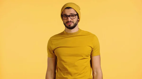 Displeased young man in beanie hat, glasses and t-shirt isolated on yellow — Fotografia de Stock