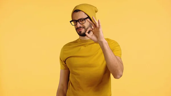 Молодой человек в шапочке, очках и футболке показывает хорошо изолированный на желтом — стоковое фото
