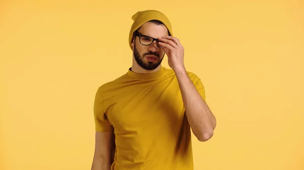 Young man in beanie hat puffing cheeks and adjusting glasses isolated on yellow — Stock Photo